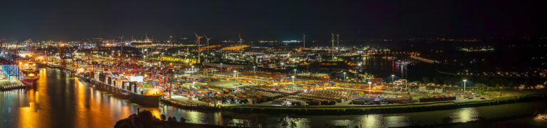 The picture shows the bustling Port of Hamburg at night, with numerous container terminals, cranes and ships. The site is dotted with many lights, which emphasise its industrial character. Large container gantry cranes can be seen at the Eurogate Terminal, with red supports and blue crossbeams, as well as numerous other cranes and storage areas for container units. The waterfront is visible, with several ships moored there, including a CMA CGM container ship. The entire scene is brought to life by the facility’s lighting and the reflections on the water.