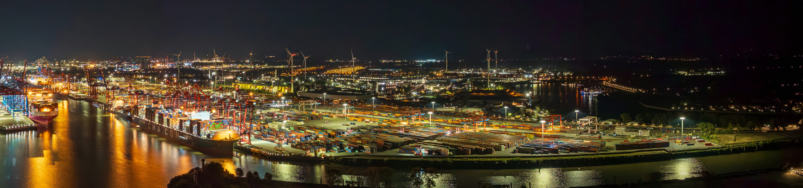 The picture shows the bustling Port of Hamburg at night, with numerous container terminals, cranes and ships. The site is dotted with many lights, which emphasise its industrial character. Large container gantry cranes can be seen at the Eurogate Terminal, with red supports and blue crossbeams, as well as numerous other cranes and storage areas for container units. The waterfront is visible, with several ships moored there, including a CMA CGM container ship. The entire scene is brought to life by the facility’s lighting and the reflections on the water.